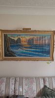 Full view of large framed oil painting of coastal landscape with waves and cliffs, illuminated by attached lamp.