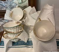 Stack of bone china teacups with gold floral pattern and gold handles on a cloth surface with packing material.