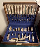 Full view of the flatware set arranged inside an open wooden case with blue velvet and cream satin lining, showing knives at the top with forks and spoons below.