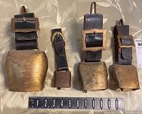Four cowbells of different sizes on wide dark brown leather straps with decorative buckles displayed on a light fabric background with a ruler for size reference.