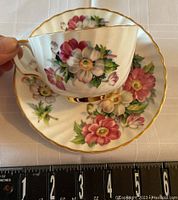 Hand-held tea cup showing floral design and gold trim with matching saucer beneath on white cloth, a ruler visible for size reference.
