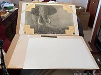 Print displayed on table, front view showing black and white elephant image, mounted to backing with corner tape, cardboard backing visible.