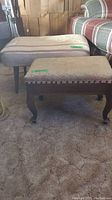 Two upholstered foot stools on carpeted floor, one larger with straight legs, one smaller with curved legs and nailhead trim, showing wear.