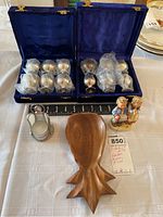 Two sets of six silver-colored metal goblets in blue velvet cases, wooden pineapple tray, angel votive holder, and Goebel figurine on table.