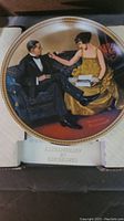 Porcelain collector plate showing a man in a tuxedo and a woman in a gold dress seated on a couch, with certificate underneath visible