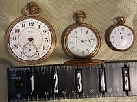 Three pocket watches placed next to a measuring tape showing size; includes Elgin and two other watches, one hunter-style.