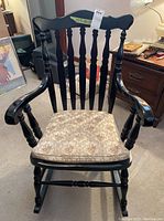 Front view of dark finished wooden rocking chair with floral patterned fabric cushion on seat, curved armrests, vertical spindle backrest with scalloped top rail.