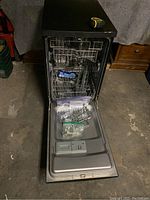 Front view of open dishwasher showing interior racks and door with detergent dispenser. Additional hardware bags placed inside the unit.
