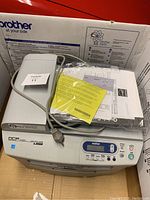 Front angled view of Brother DCP-7020 laser printer on floor with power cord and manuals on top, showing control panel and top scanner lid.
