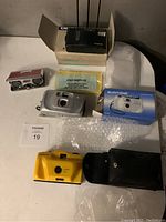 Three new cameras displayed on white surface: Olympus Infinity Junior, Suntone MM35E, and yellow camera with black carrying case. Also includes bubble wrap and manual.