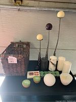 Photo showing candle lot items on a black surface including a rectangular wicker basket, three tall metal candle holders with round purple and white candles, three white pillar candles, two green and white round candles, a pack of green tea lights, a large textured white round candle, and a bowl-shaped candle.