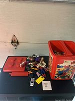 Photo of the red Lego bin next to the lid and various small toys including cars on a black surface.