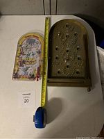 Two tabletop pinball games side by side on a table with measuring tape showing size. One has a Curious George colorful plastic design, the other is wooden with metal pins.