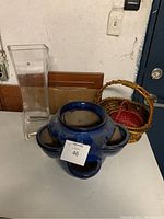 Photo showing tall clear glass vase, blue ceramic flower pot with four side pockets, and two small wicker baskets on a white surface.
