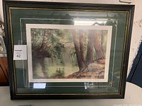 Full view of the framed art print showing the riverside scene with trees and water reflections in a black frame with gold accents and a double mat.