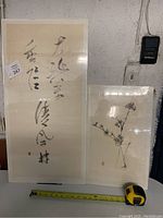 Two unframed rice paper paintings: one calligraphy, one floral vase sketch, shown with measuring tape.