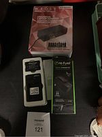 Photo showing the KROSS universal laptop adapter box with product and multiple tips displayed, alongside Refuel travel charger kit box and Canon battery with charger out of packaging.