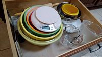 Drawer with stacked assorted bowls in yellow, green, orange, and white, a wooden rolling pin, Camry white digital kitchen scale with round top, glass measuring cup, Pyrex glass bowl, metal bowls, and a Tupperware bowl without lid.