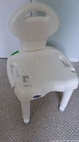 Front profile of white plastic bath seat showing perforated seat surface, arm handles, backrest with handle cutout.