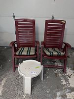 Two red plastic folding chairs each with a striped beige, green, and white seat cushion, placed next to a small white plastic side table on a concrete floor.
