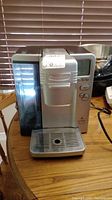 Front view of the silver Cuisinart Keurig coffee maker with water reservoir and drip tray on a wooden table.