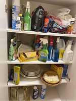White cabinet shelf holding household cleaning products, some plates, a basket with napkins, and kitchen/dining items