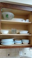 Wood cabinet shelf with stacks of white dishes including plates, bowls, and saucers, plus small white ramekins and large glass bowls.
