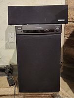 Kenwood 103SW powered subwoofer with Energy Shielded ECC-1R center channel speaker on top placed against a wall with power outlet visible. Remote is on the floor next to speaker.