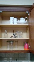 Wooden kitchen cabinet with white Sadler teapot, two white Corelle coffee cups, glass bottle with dropper, corkscrew, glasses, and red kitchen item.