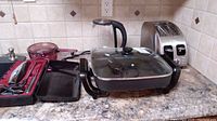 Kitchen countertop displaying all appliances and utensils in the lot including electric skillet with glass lid, electric kettle, toaster, glass bowl, and electric cutting utensils in case.
