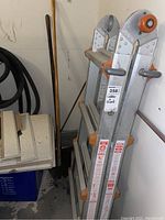 Aluminum folding ladder standing upright with two wooden handled garden tools behind it.