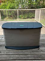 Front view of a large Rubbermaid outdoor storage box with a closed dark green lid and beige body on a wooden deck.