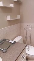 Bathroom corner showing Weight Watchers clear glass digital scale on countertop, metal freestanding toilet paper holder, and three small white wood wall shelves mounted on beige wall.