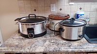 Three slow cookers on a kitchen counter including Bravetti stainless steel slow cooker, Hamilton Beach decorated ceramic slow cooker, and Rival stainless steel slow cooker.