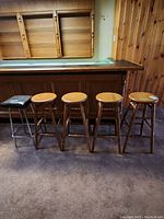 Set of 5 bar stools in a basement setting. Four matching oak wood stools with round tops and traditional turned legs. One metal frame stool with black faux leather upholstery on seat and chrome legs, all positioned in front of wooden paneled wall and bar counter.