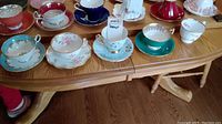 Wide angle photo showing thirteen assorted teacups and saucers featuring various floral and solid colored ceramic designs, arranged on a wooden table.
