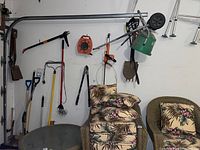 Green seed spreader with metal frame laying on the wall adjacent to other garden tools like rakes and shovels. Some wicker chairs with floral cushions are also visible.