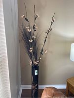 Tall dark wood vase with carved floral designs on the floor next to a chair and lamp, containing tall faux flower stems and decorative branches.