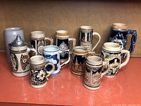 Photo showing nine assorted ceramic beer steins in various sizes and designs on an orange surface, displaying the full collection.