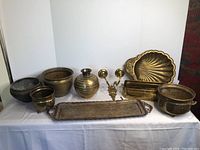 Full view of assorted copper and brass items arranged on white cloth backdrop showing trays, pots, and decorative candle holder.