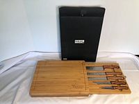 Full view of wooden cutting board with sliding compartment open showing five knives. Board engraved with 'Doctors Nova Scotia'. Black carrying/storage bag labeled 'Knife Set & Cutting Board' in background.