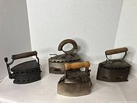 Four vintage charcoal irons arranged in two rows showing wooden handles and overall metal bodies with patina and wear.