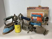 Three vintage Coleman irons with fuel tanks and handles along with a measuring can and flat metal tool, positioned together to show full set