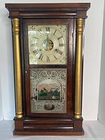 Front view of the vintage wooden ogee clock. Visible painted glass panel on lower door featuring a landscape scene and gold-tone columns on sides.