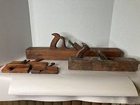 Front view of two wooden wood planes and three wooden plane accessories on white surface with neutral backdrop.