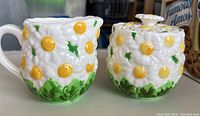 Creamer and sugar bowl set with yellow-centered daisies and green leaves, textured floral design, placed on table.