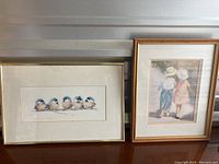 Two framed art prints side by side on a shelf: one horizontal with birds, one vertical with children