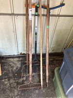Photo showing six garden tools standing against a wall, including two rakes, two hand cultivators, one metal hoe, and one three-prong pitch fork with wooden handles.