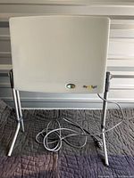 Front view of the white rectangular daylight lamp on its silver metal stand with power cord coiled in front.