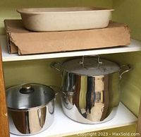View of three items stacked on shelves: a beige rectangular ceramic stone baker on top shelf, and two shiny stainless steel pots with lids on the lower shelf, showing their sizes and shapes.